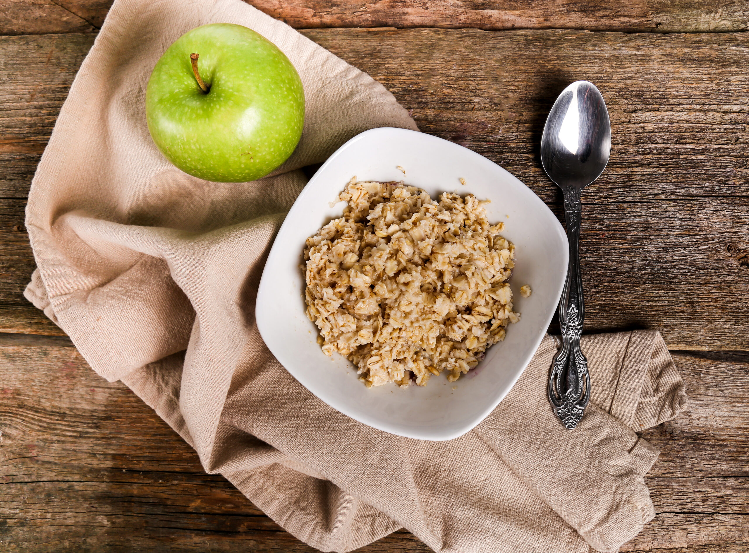 A bowl of plain oatmeal with a green apple and a spoon on a rustic wooden table.